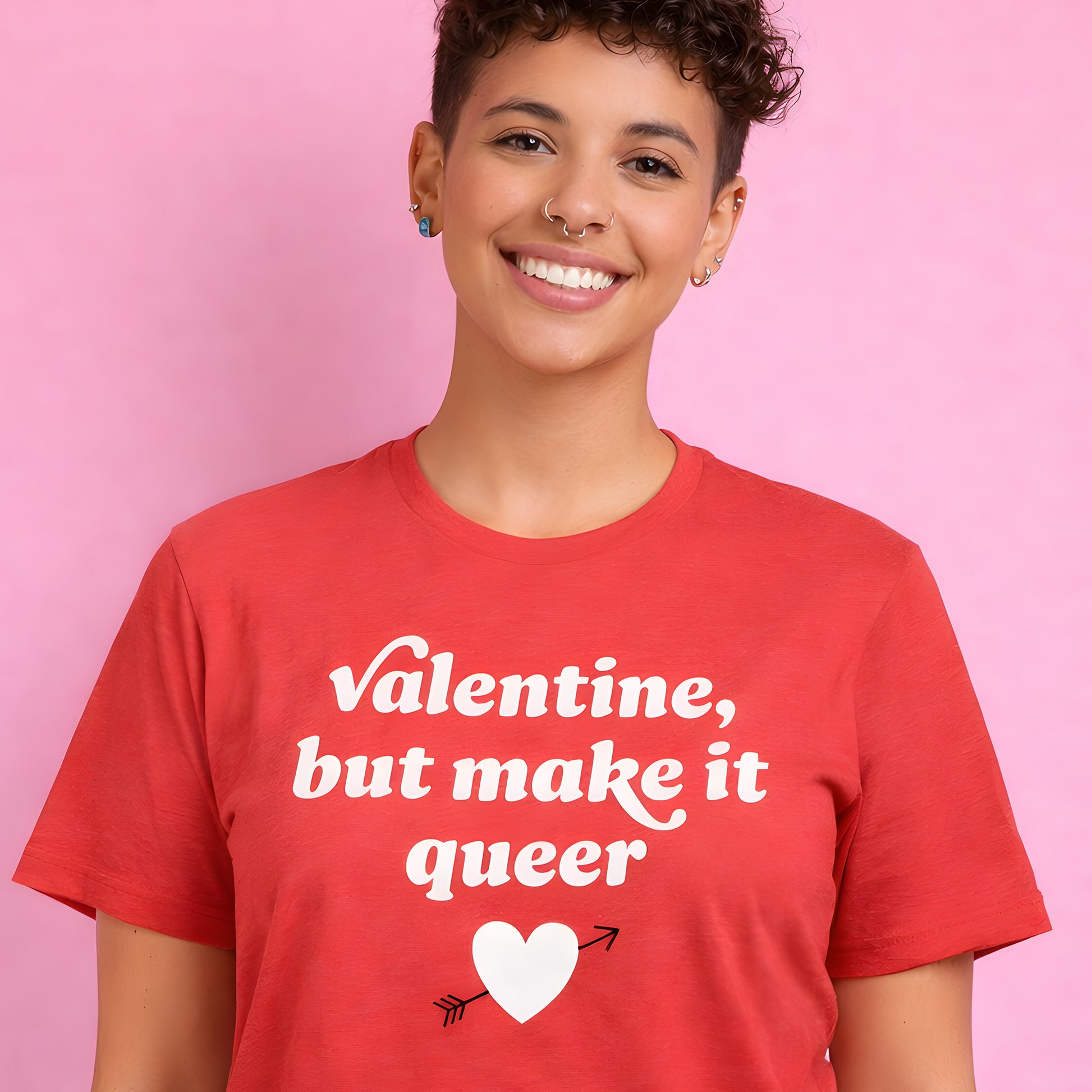 Unisex heather red t-shirt worn by a smiling model featuring bold retro typography and heart graphic with the phrase “Valentine, but make it queer,” a progressive Valentine’s Day design.