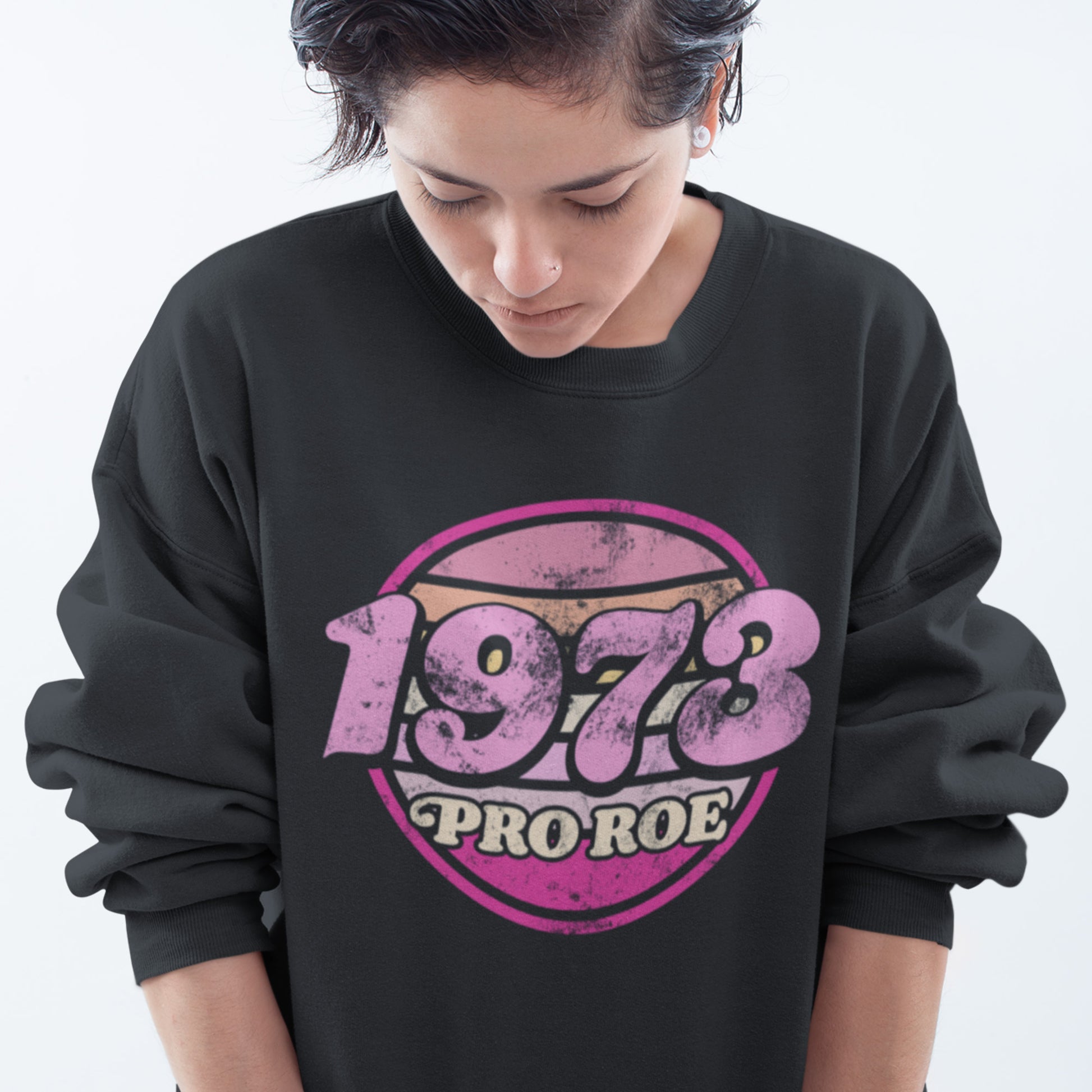 Model wearing a black unisex premium sweatshirt with a faded pink circular “1973 PRO ROE” graphic, casual downward pose highlighting the centered vintage print from Blue Wave USA.