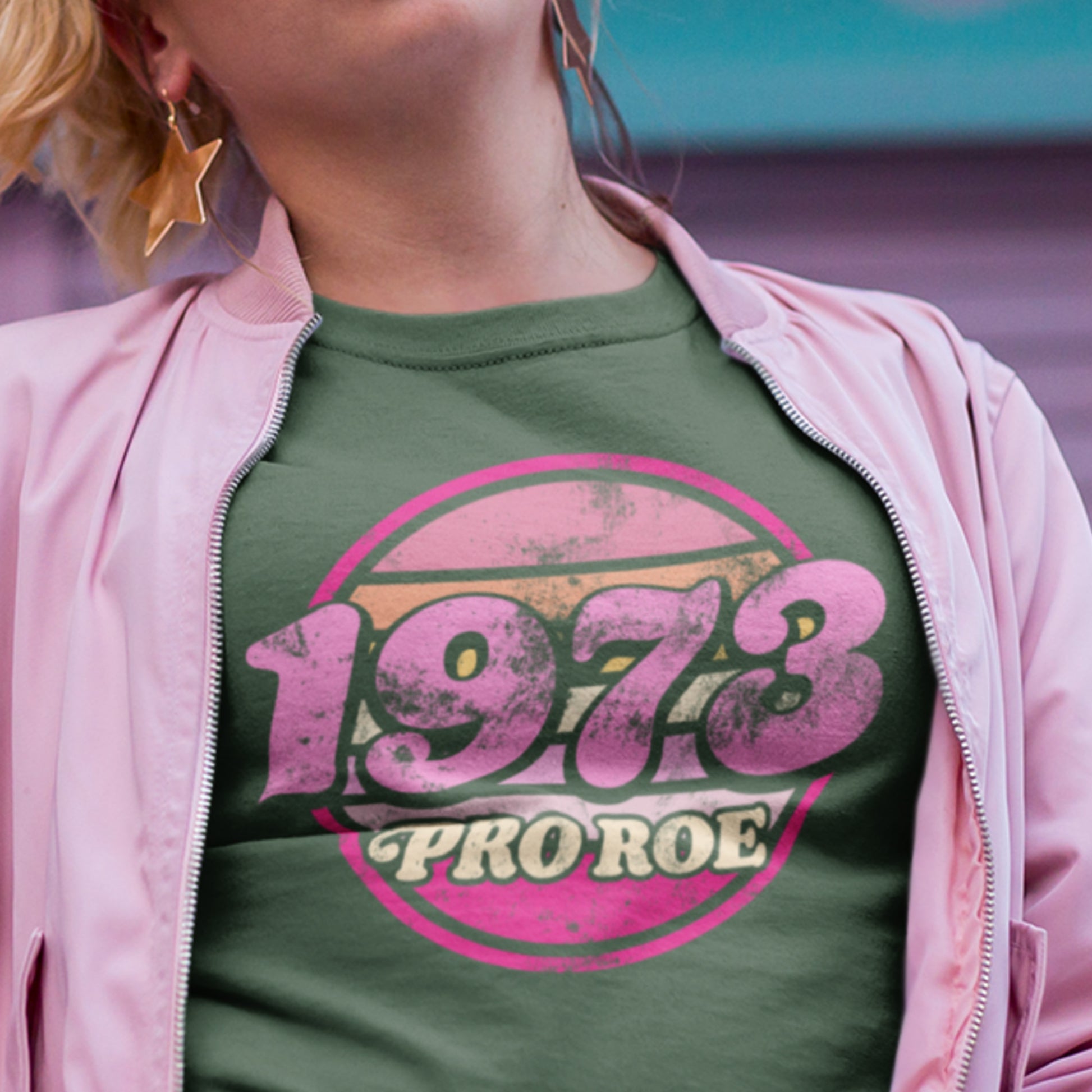 Model wearing a forest green unisex t-shirt with a retro circular “1973 PRO ROE” graphic in pink tones, styled casually in an outdoor setting from Blue Wave USA.