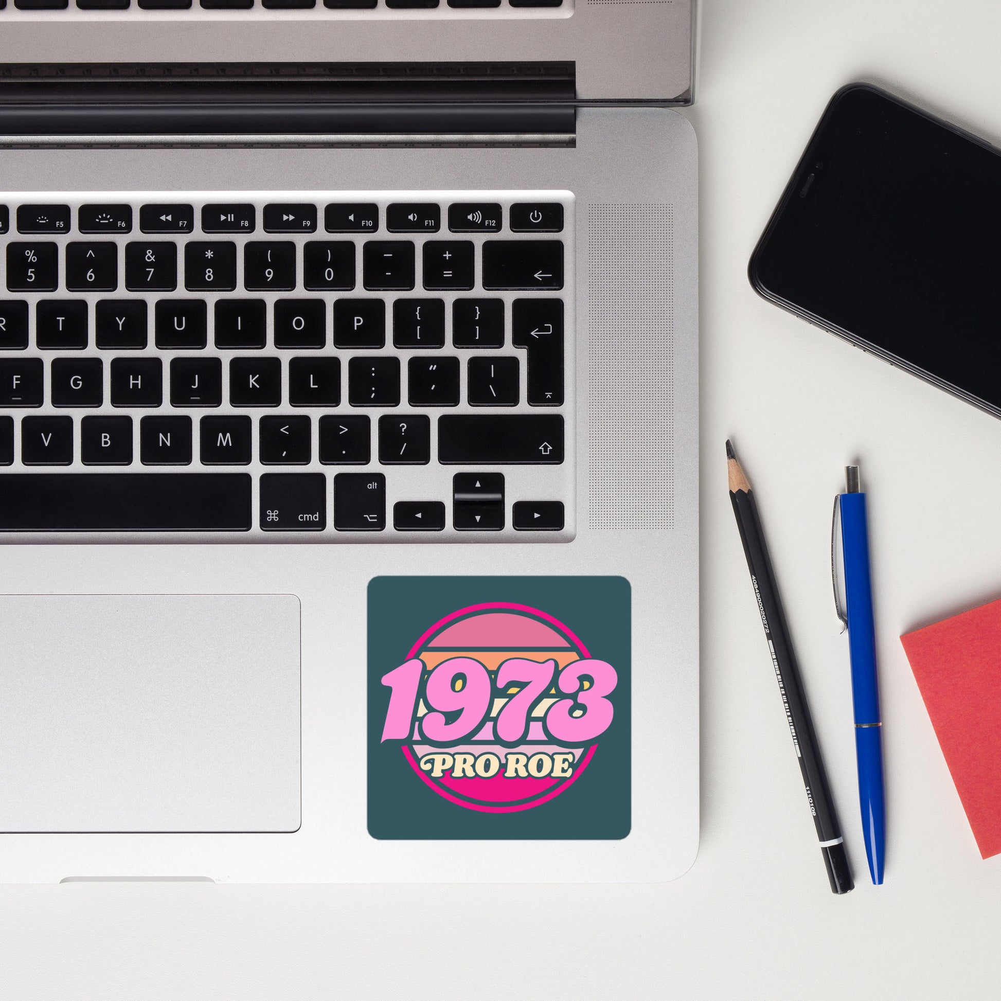 Vintage “1973 PRO ROE” sticker shown on a laptop beside notebooks and stationery, highlighting the retro pink circular badge design as reproductive freedom sticker from Blue Wave USA.
