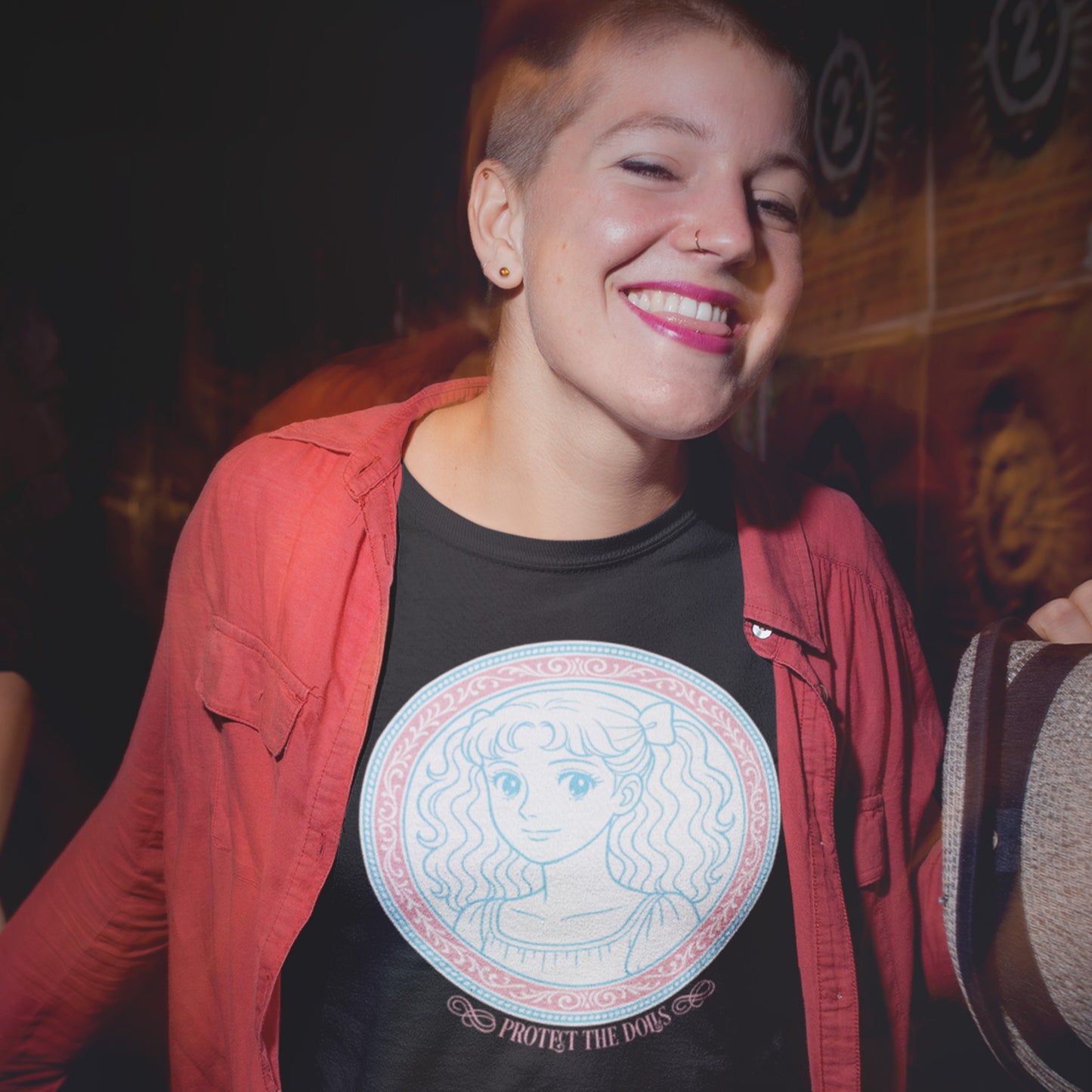 Person smiling while wearing the “Protect the Dolls” t-shirt layered under a red jacket, showing the soft pink-and-blue artwork that celebrates trans femme strength and pride.