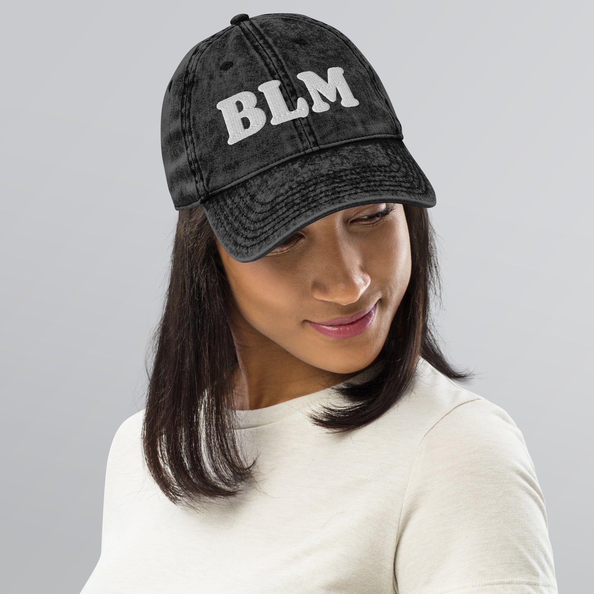 Model wearing a black vintage cotton twill cap with raised embroidered “BLM” lettering, curved brim, and washed fabric texture in a neutral studio setting.