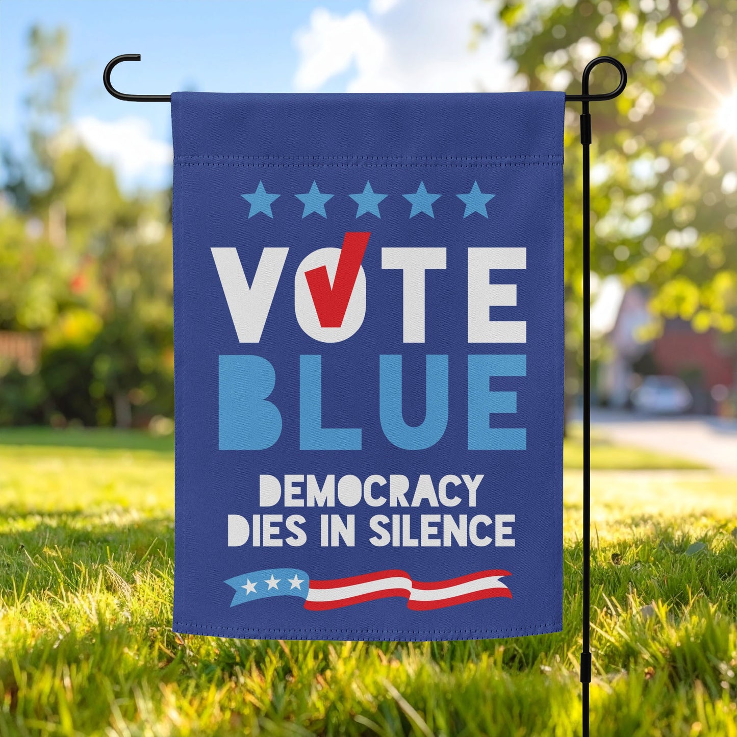 A "Vote Blue: Democracy Dies in Silence" garden flag displayed on a black metal stand in a sunny, green front yard with a blurred house background.