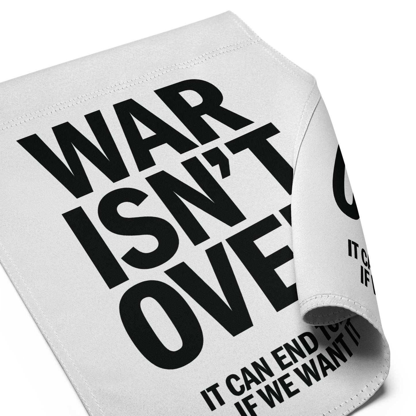 Close-up folded garden flag corner showing oversized “WAR ISN’T OVER” typography with visible “IT CAN END TODAY IF WE WANT IT” text.