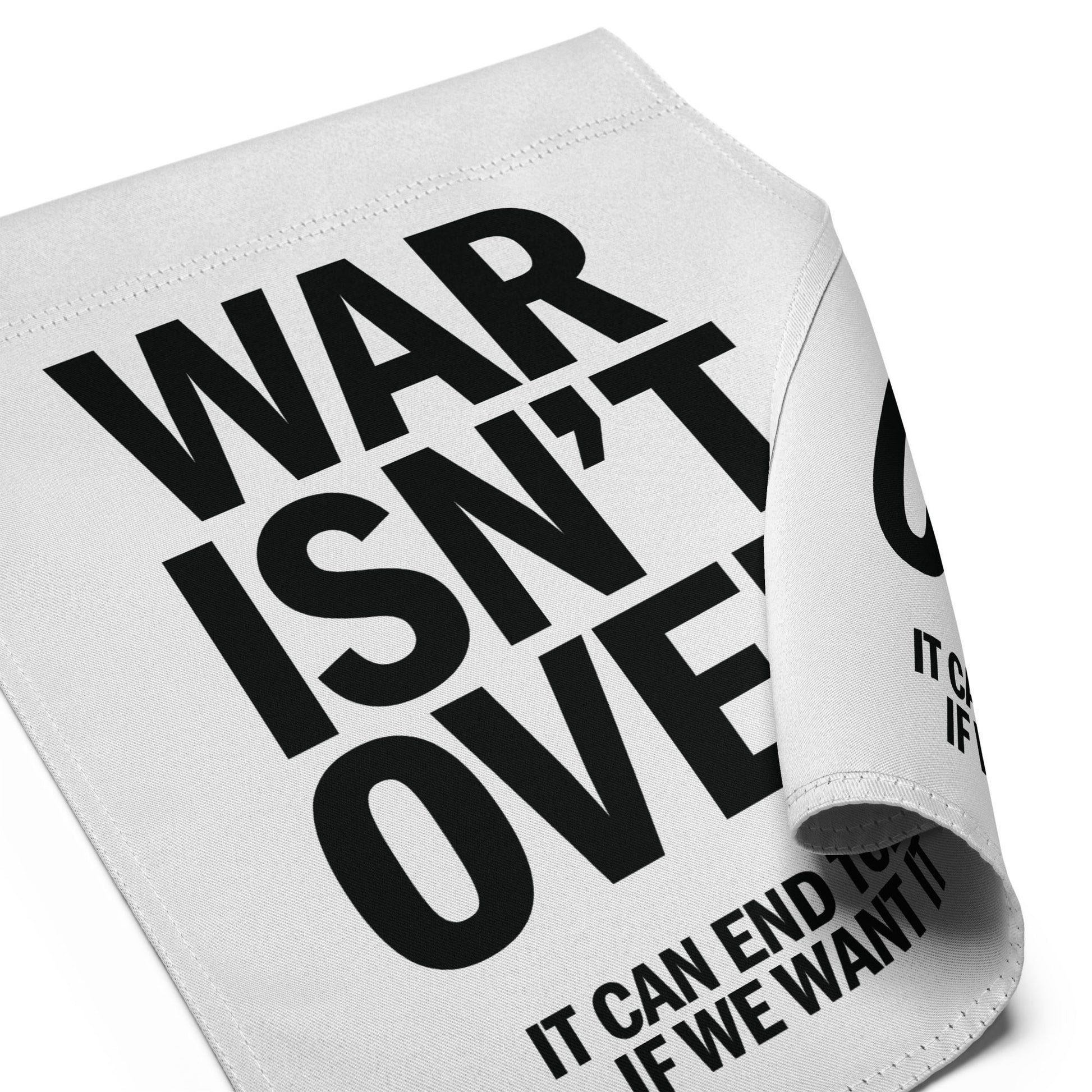 Close-up folded garden flag corner showing oversized “WAR ISN’T OVER” typography with visible “IT CAN END TODAY IF WE WANT IT” text.