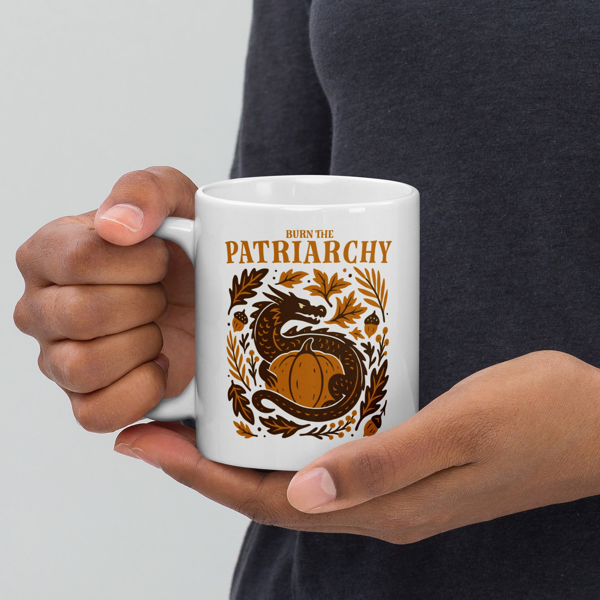 White ceramic mug with “Burn the Patriarchy” dragon and pumpkin design, feminist Thanksgiving drinkware with fall leaves and acorns.