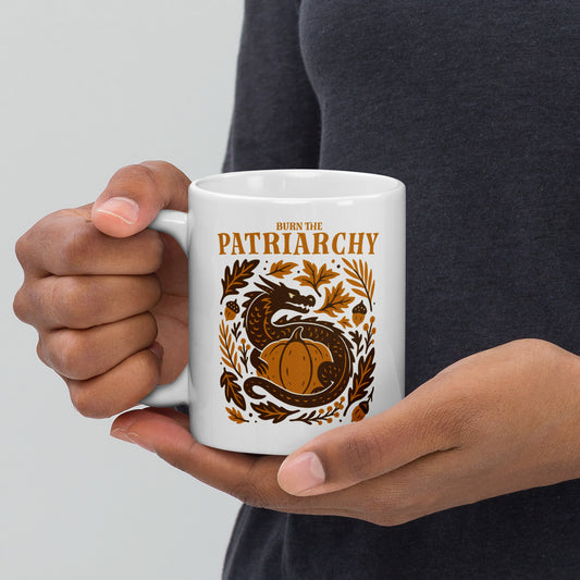White ceramic mug with “Burn the Patriarchy” dragon and pumpkin design, feminist Thanksgiving drinkware with fall leaves and acorns.