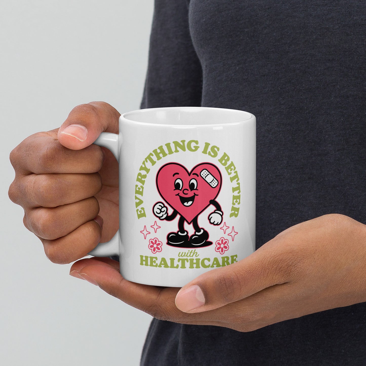 Close-up of a 11 oz white mug held in both hands, featuring a smiling pink heart character with medical icons and text that reads “Everything Is Better with Healthcare.”