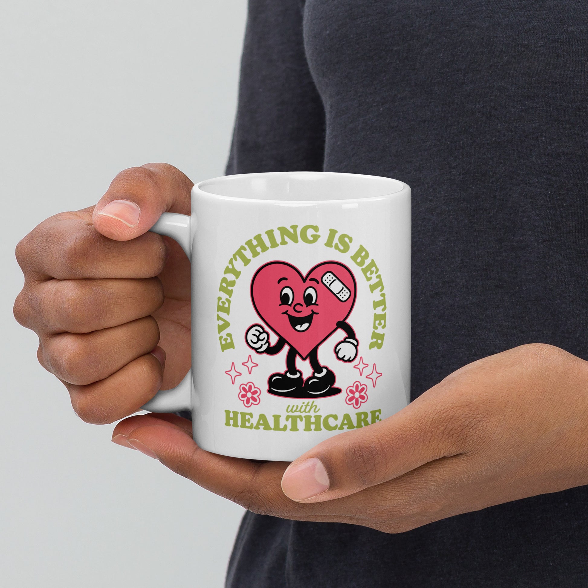 Close-up of a 11 oz white mug held in both hands, featuring a smiling pink heart character with medical icons and text that reads “Everything Is Better with Healthcare.”