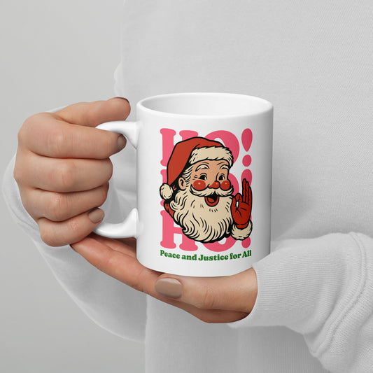 Hands holding a 11 oz white ceramic mug featuring a retro Santa illustration with pink “HO!” lettering and the printed message “Peace and Justice for All.”