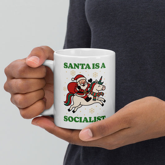 White 11 oz ceramic mug held in both hands featuring a retro Santa riding a unicorn with holiday stars and the printed message “SANTA IS A SOCIALIST.”
