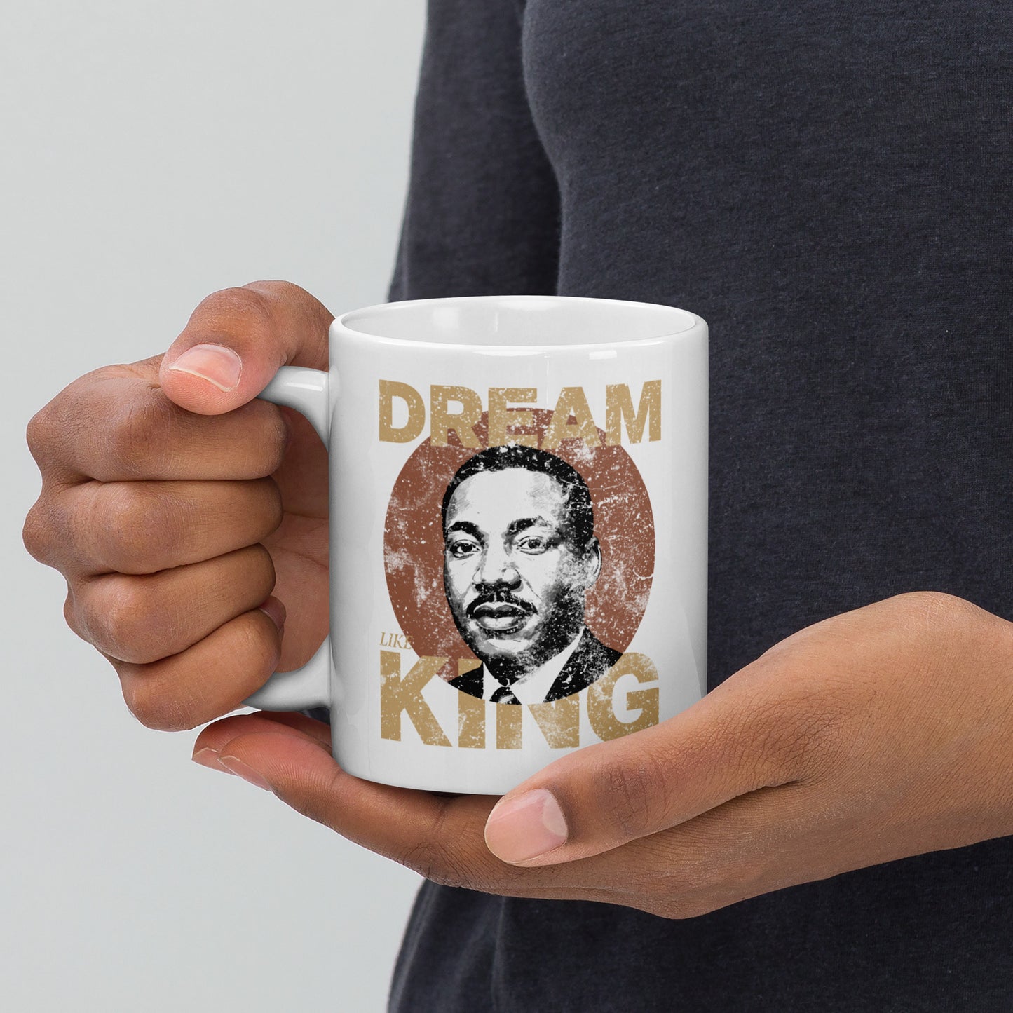 Hands holding a white 11 oz  ceramic mug printed with the distressed “DREAM LIKE KING” design featuring Martin Luther King Jr.’s portrait inside a vintage textured circle. Inspirational civil-rights style artwork.