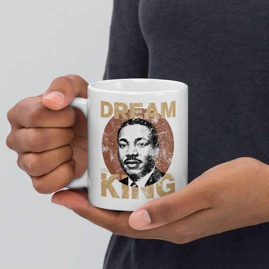 Hands holding a white 11 oz  ceramic mug printed with the distressed “DREAM LIKE KING” design featuring Martin Luther King Jr.’s portrait inside a vintage textured circle. Inspirational civil-rights style artwork.