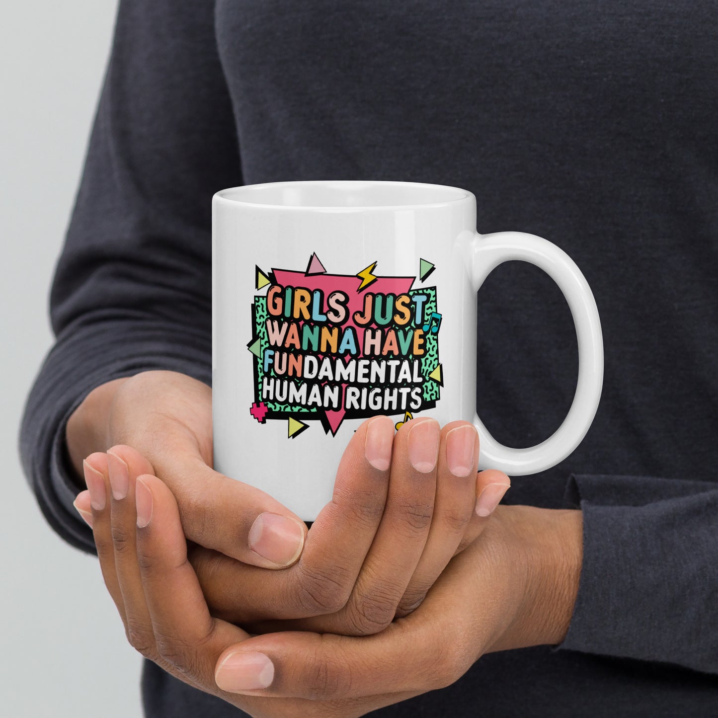 Person wearing a dark gray sweater holding the 11 oz “Girls Just Wanna Have Fundamental Human Rights” mug with both hands. Bright geometric feminist design by Blue Wave USA.