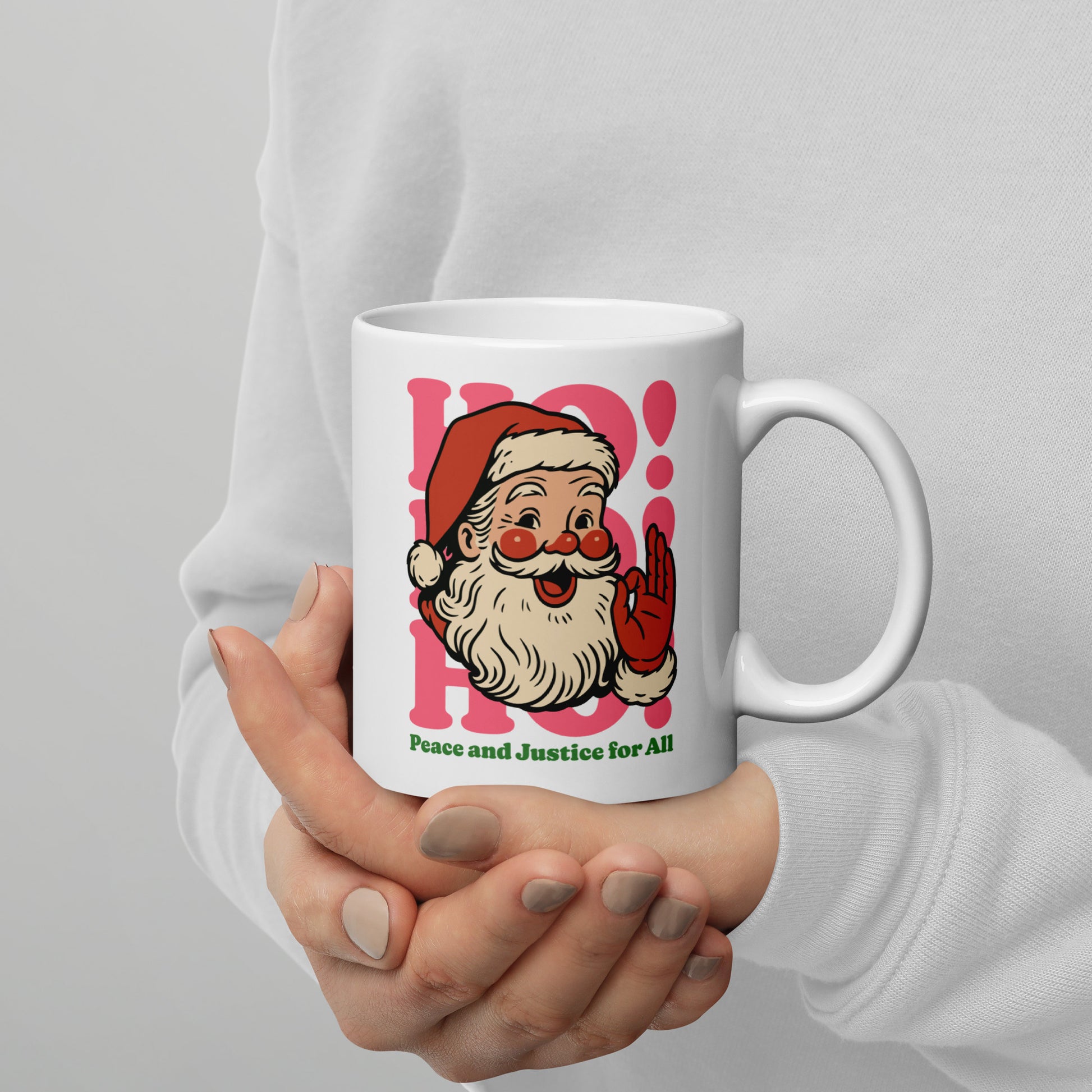 Person holding a 11 oz white ceramic mug with a vintage-style Santa graphic, pink “HO!” lettering, and the message “Peace and Justice for All,” holiday mug by Blue Wave USA.