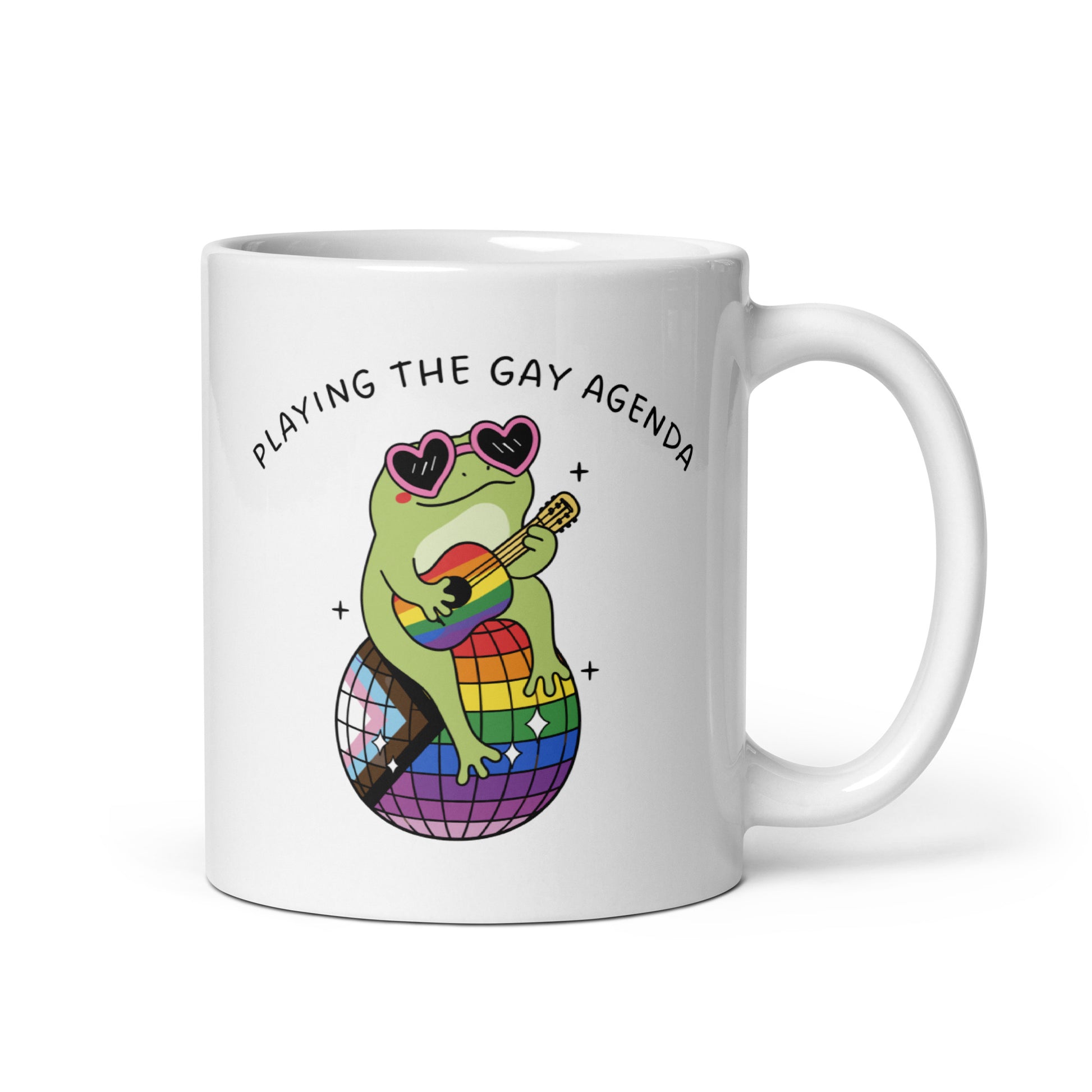 White glossy mug featuring a disco frog wearing heart-shaped sunglasses and playing a guitar atop a rainbow disco ball with “Playing the Gay Agenda” pride-inspired artwork.