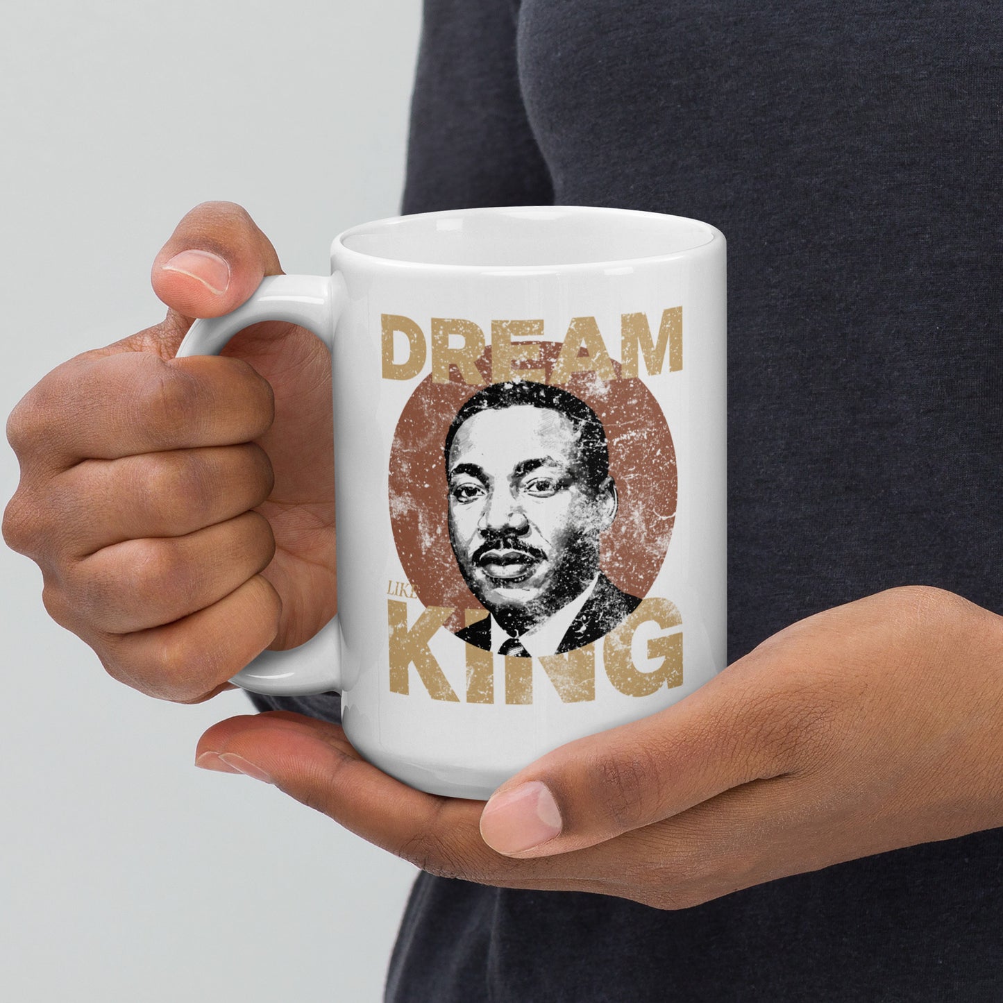Extended hands offering a white 15 oz ceramic mug featuring the distressed “DREAM LIKE KING” graphic with MLK’s portrait inside a textured circle. Retro inspirational design from Blue Wave USA.