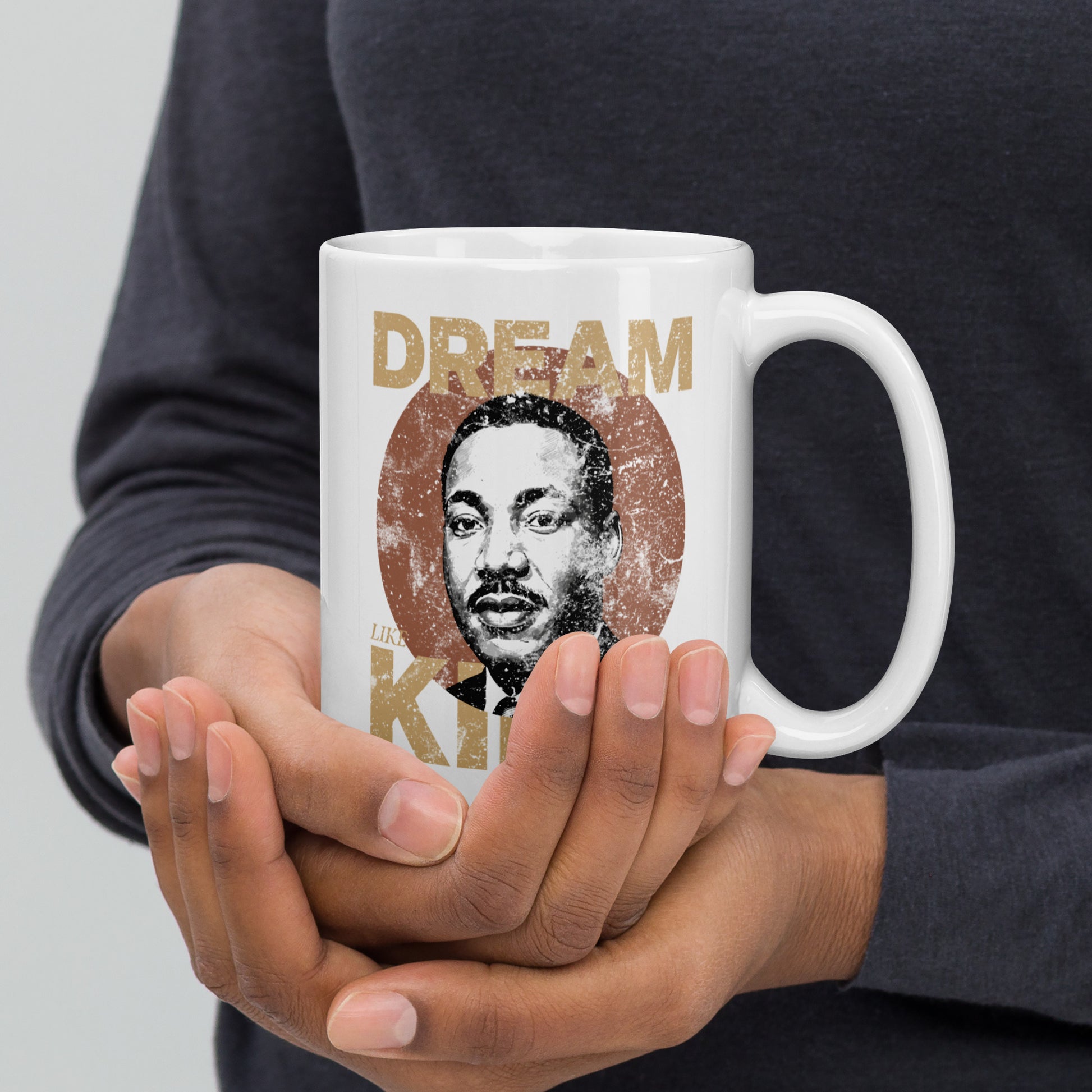 Hands gently cupping a white 15 oz mug printed with the aged “DREAM LIKE KING” lettering and Martin Luther King Jr.’s portrait in a weathered circle. Vintage civil-rights themed artwork.