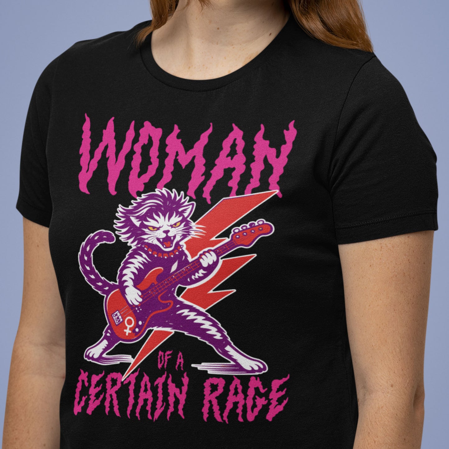 Model wearing a black unisex t-shirt with a punk rock cat electric guitar illustration and distressed text reading “WOMAN OF A CERTAIN RAGE,” casual studio lifestyle image from Blue Wave USA.