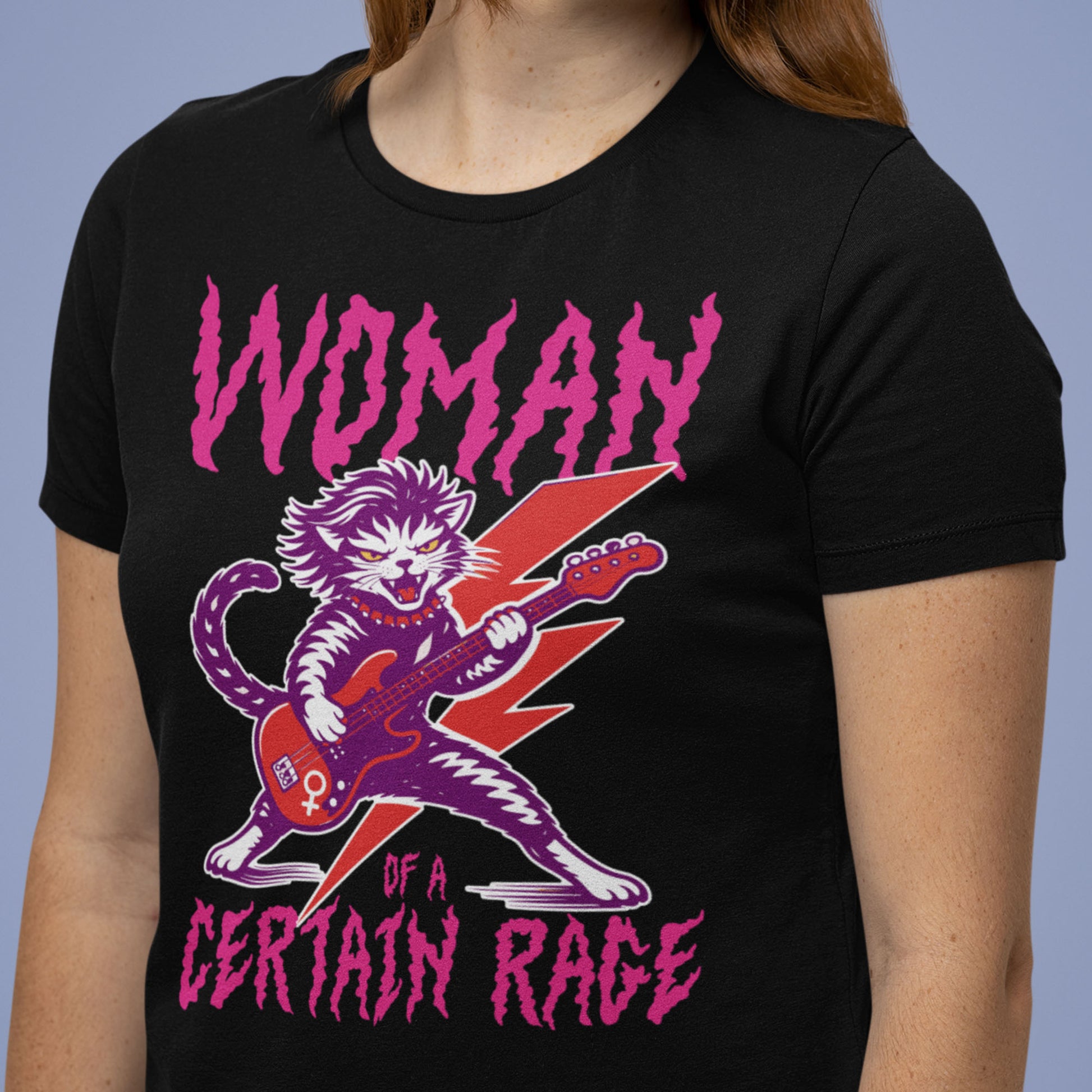 Model wearing a black unisex t-shirt with a punk rock cat electric guitar illustration and distressed text reading “WOMAN OF A CERTAIN RAGE,” casual studio lifestyle image from Blue Wave USA.