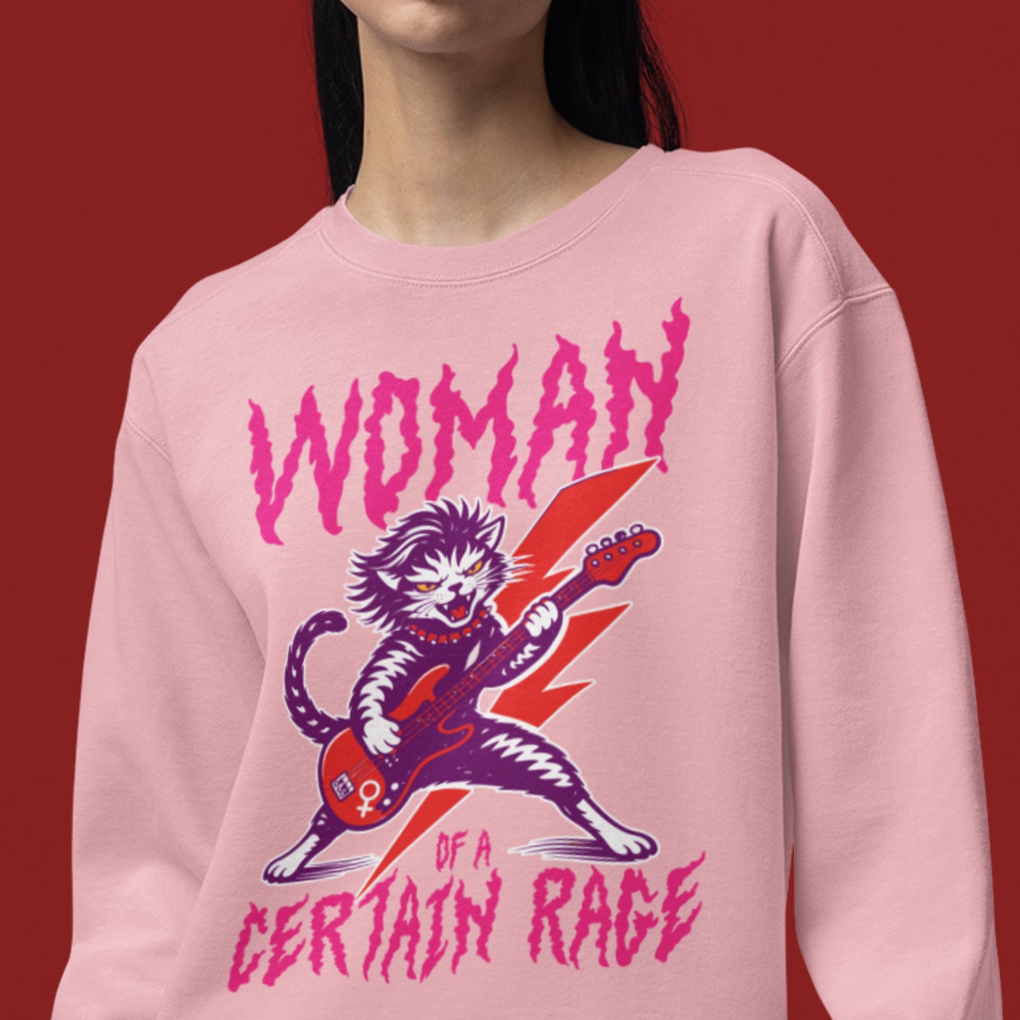 Model wearing a light pink unisex premium sweatshirt with a feminist punk rock cat electric guitar illustration and distressed text reading “WOMAN OF A CERTAIN RAGE,” studio lifestyle image from Blue Wave USA.