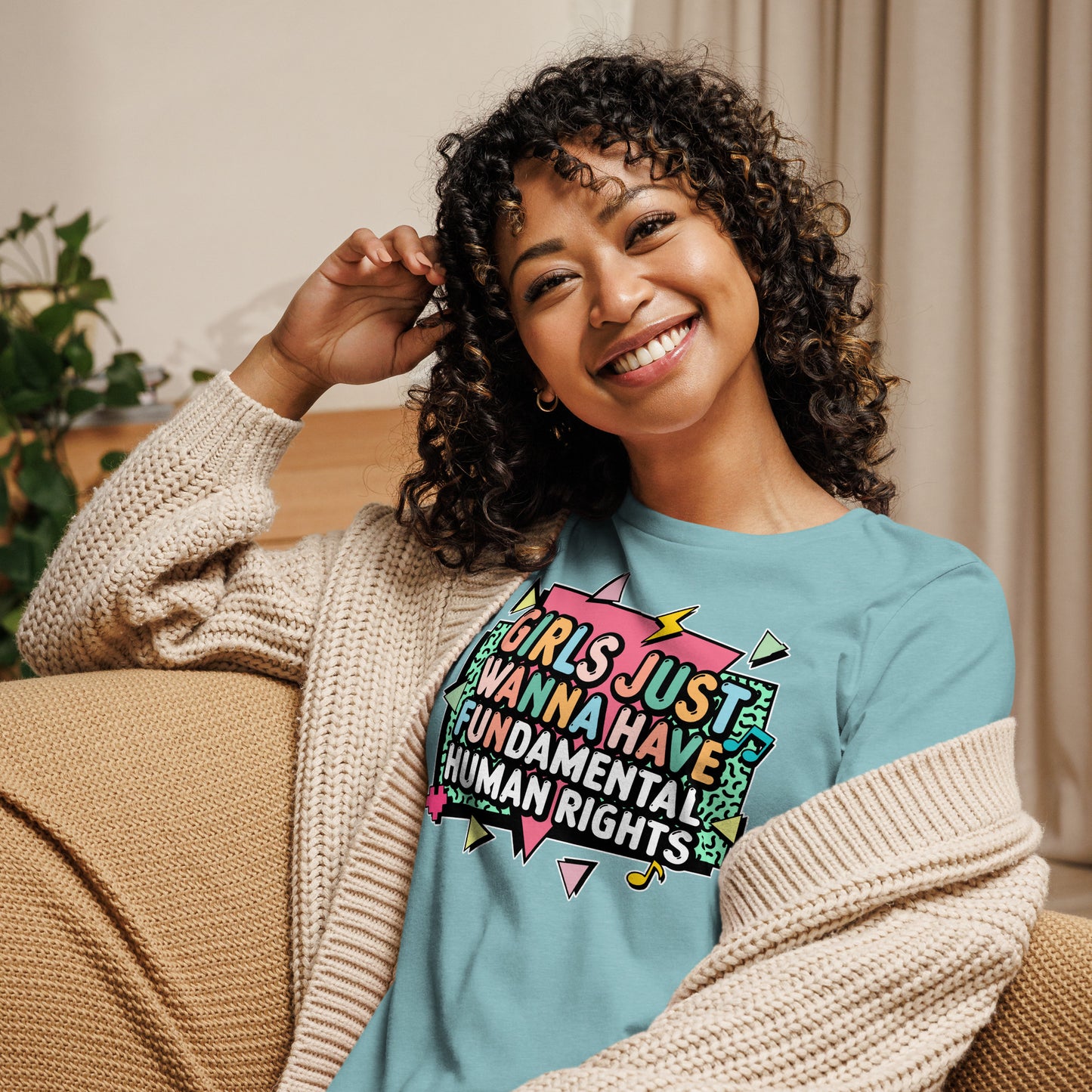 Model relaxing on a sofa wearing the Heather Blue Lagoon “Girls Just Wanna Have Fundamental Human Rights” t-shirt with pastel shapes and bold lettering.