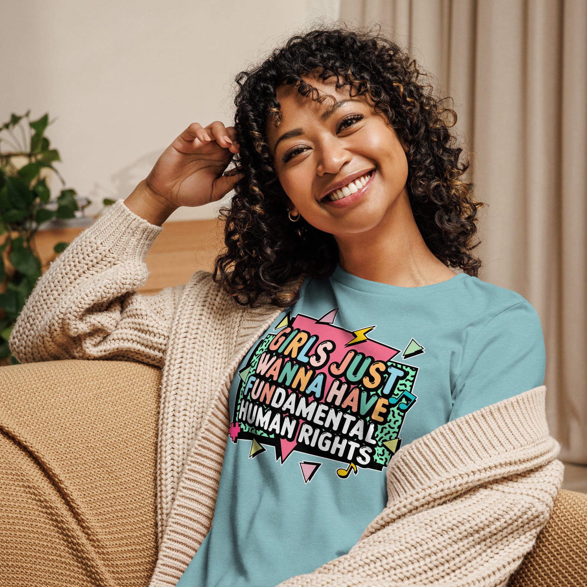 Model relaxing on a sofa wearing the Heather Blue Lagoon “Girls Just Wanna Have Fundamental Human Rights” t-shirt with pastel shapes and bold lettering.
