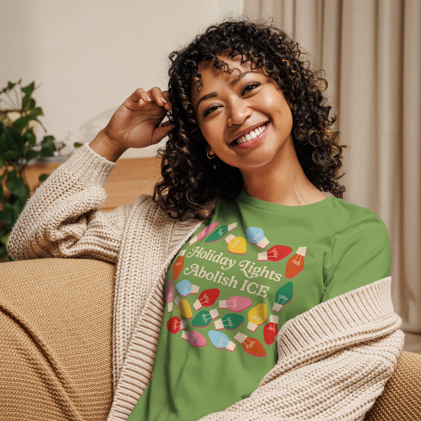 A relaxed model sits on a couch wearing a leaf green Women’s Relaxed T-Shirt featuring holiday bulbs framing the message “Holiday Lights Abolish ICE,” styled for cozy seasonal activism from Blue Wave USA.
