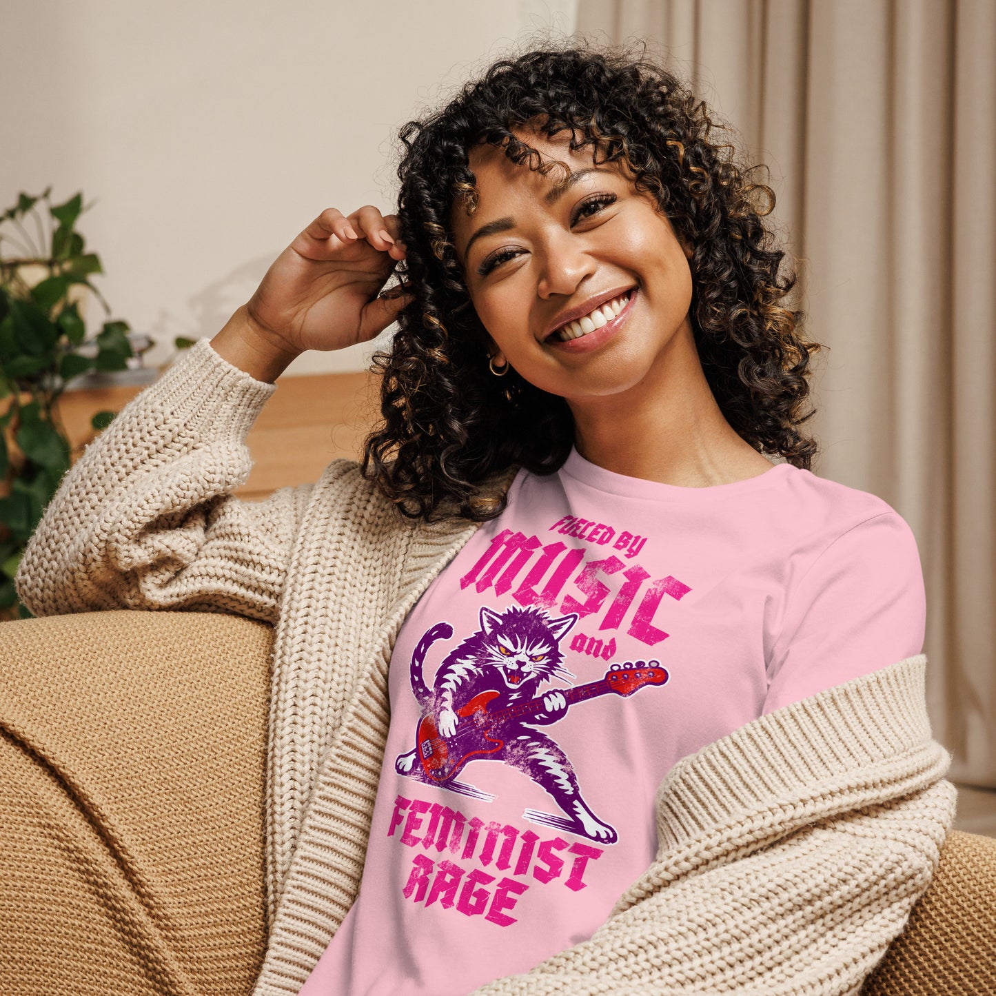 Model wearing a light pink women’s relaxed t-shirt with a punk rock cat guitar illustration and the phrase “FUELED BY MUSIC AND FEMINIST RAGE,” casual indoor lifestyle setting.
