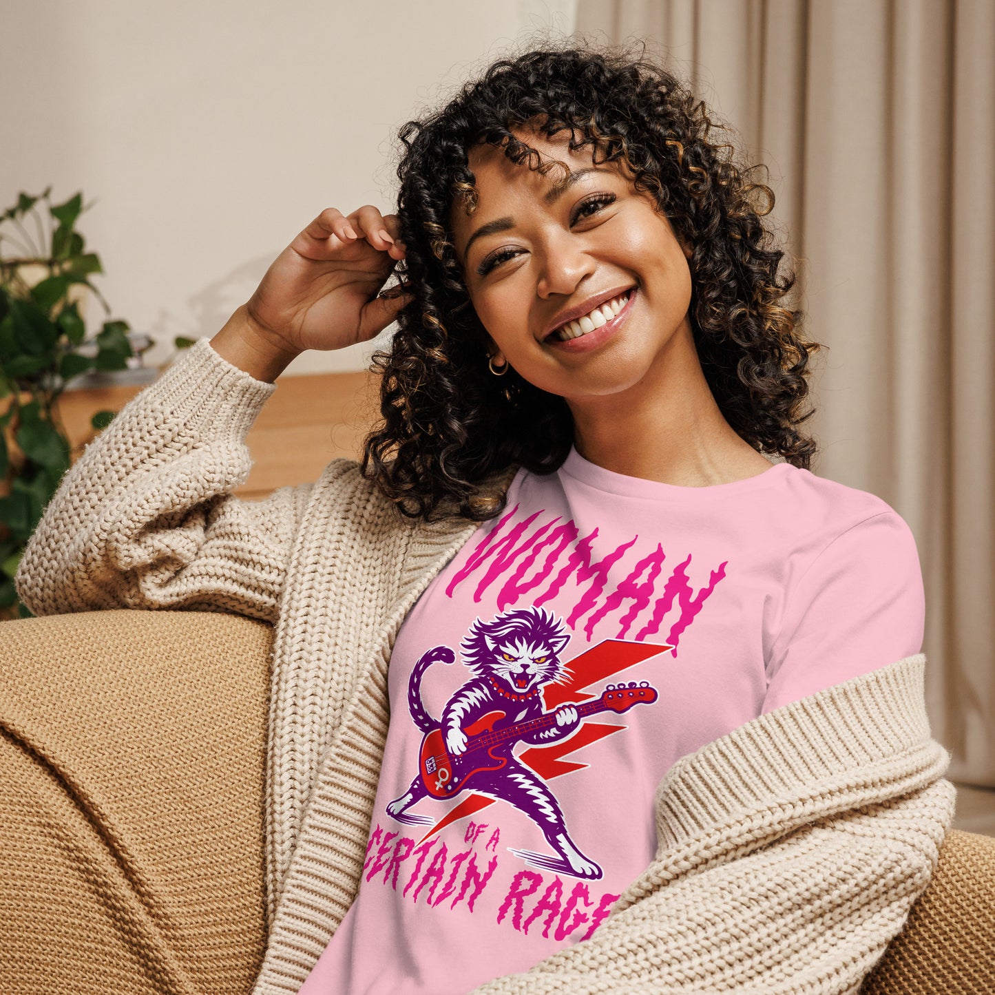 Model wearing a light pink women’s relaxed t-shirt with a feminist punk rock cat electric guitar illustration and distressed lettering reading “WOMAN OF A CERTAIN RAGE,” cozy indoor setting.