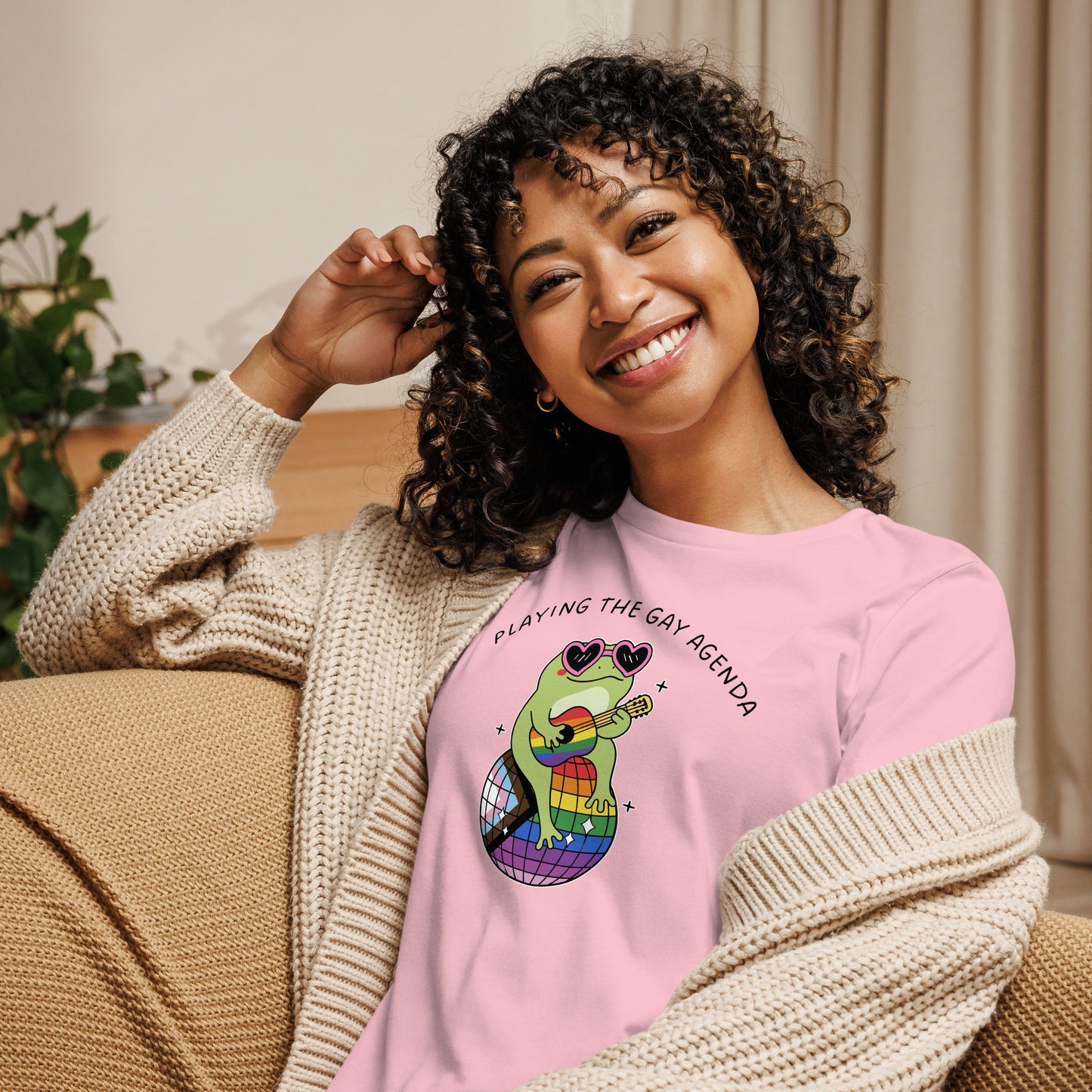 Woman wearing a pink pride t-shirt featuring a disco frog playing guitar atop a rainbow disco ball with playful “Playing the Gay Agenda” artwork.