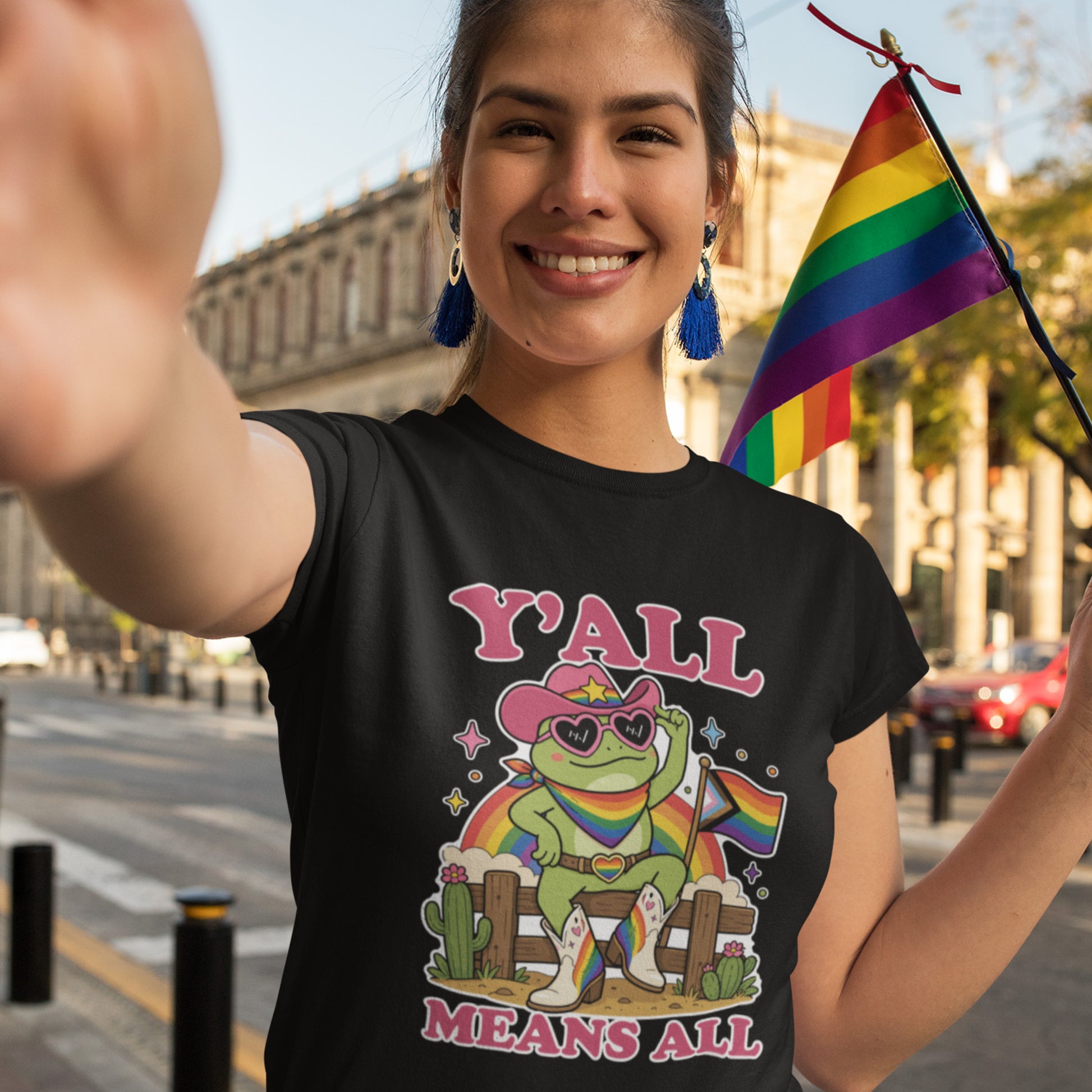 Person wearing a black unisex t-shirt featuring a colorful cowboy frog in heart-shaped sunglasses holding a rainbow Pride flag alongside the phrase “Y’All Means All,” photographed outdoors during a Pride celebration.