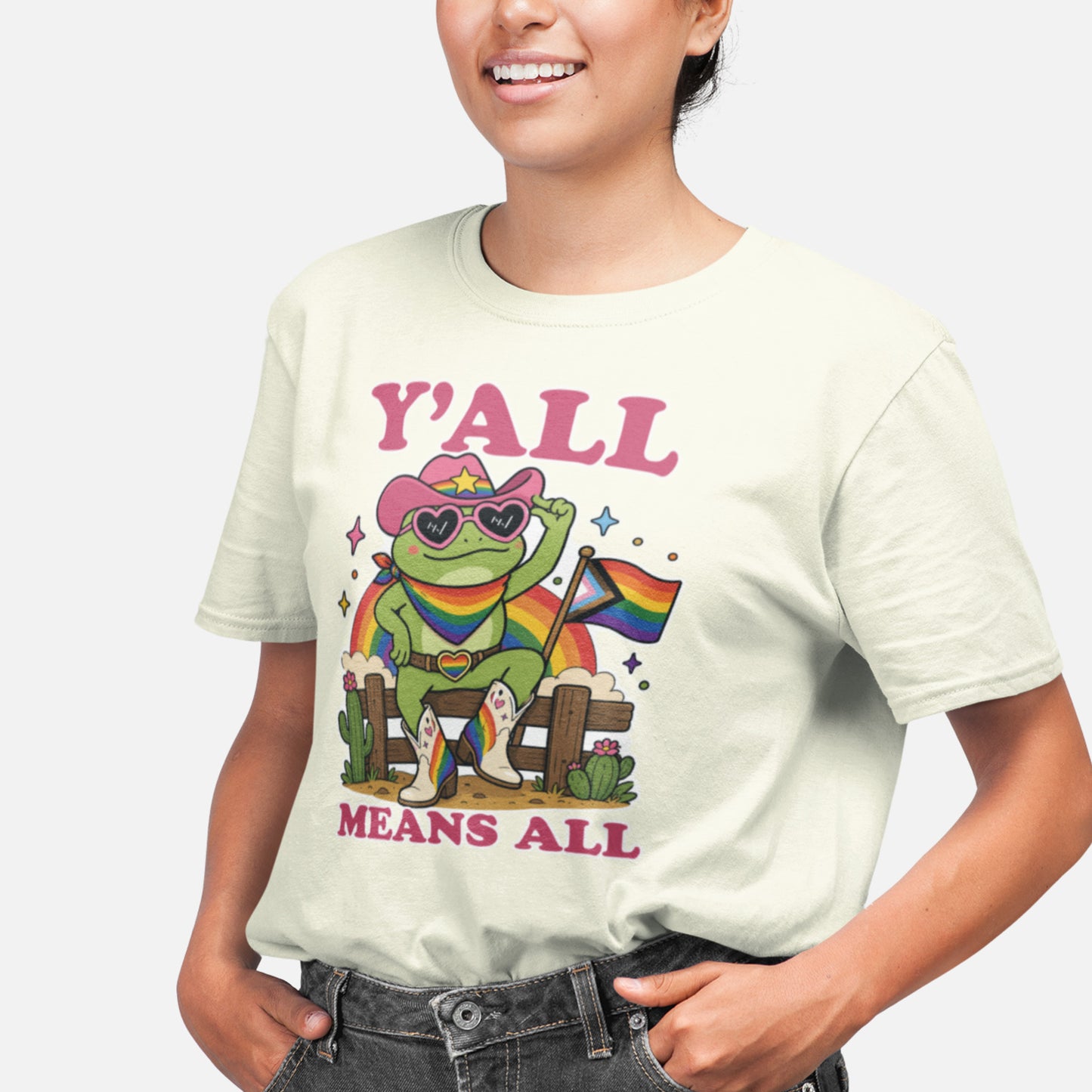 Person wearing a cream-colored unisex t-shirt featuring a rainbow cowboy frog graphic with Pride flag, western-inspired artwork, and the inclusive message “Y’All Means All” supporting LGBTQ+ visibility and belonging.