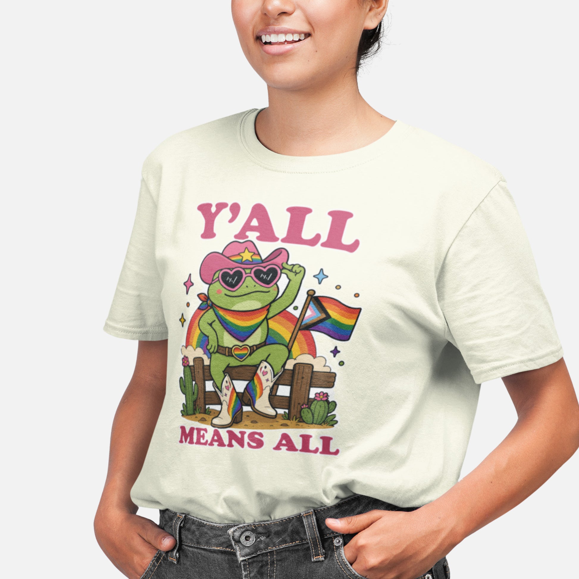 Person wearing a cream-colored unisex t-shirt featuring a rainbow cowboy frog graphic with Pride flag, western-inspired artwork, and the inclusive message “Y’All Means All” supporting LGBTQ+ visibility and belonging.
